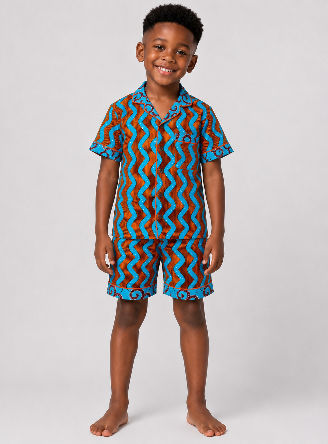 A young boy stands barefoot against a light gray studio background, wearing a matching short-sleeve button-up shirt and shorts set with a bold wavy zigzag pattern in turquoise blue, brown, and black.